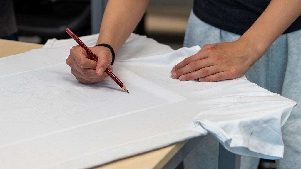 Leerling schetst met potlood op het t-shirt tijdens de Workshop T-Shirt Ontwerpen.
