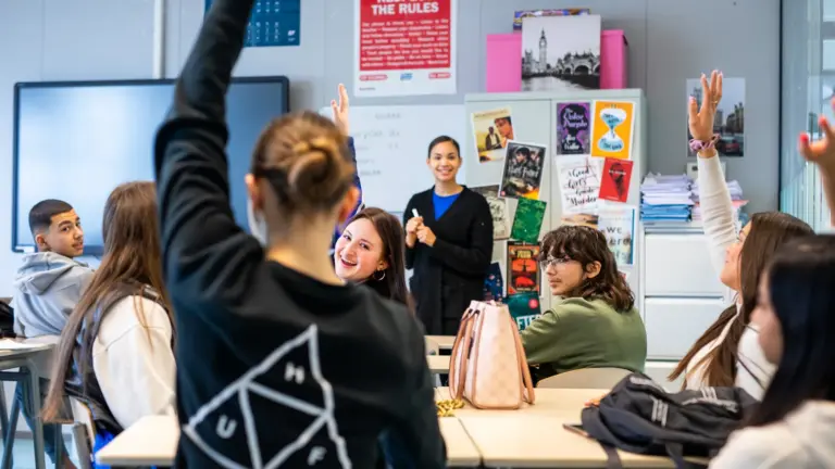 Leerlingen steken hun hand op in de klas terwijl een begeleidster voor de groep staat